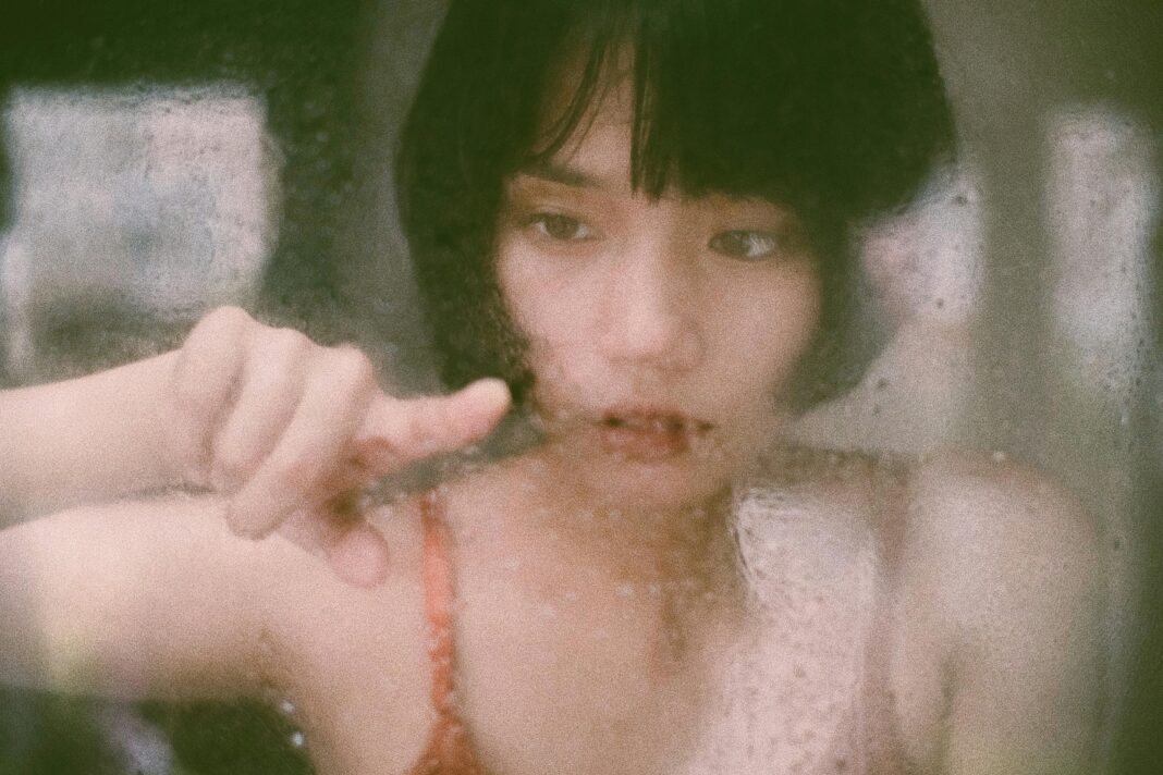 Through window of young Asian woman drawing on wet glass with finger in soft light